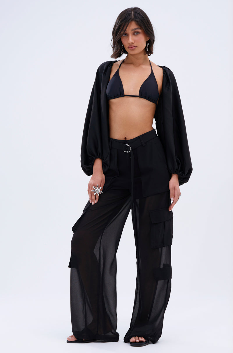 Woman wears a black bikini top, sheer black cargo pants with pockets, and the COVER UP CHIFFON SHRUG IN BLACK. She stands against a plain light background with wavy hair and accessorizes with rings.