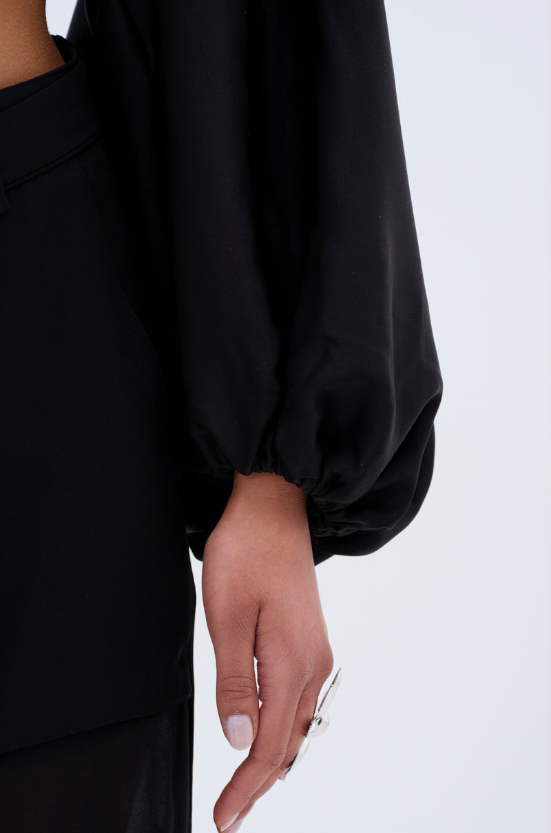 A close-up of an arm wearing the COVER UP CHIFFON SHRUG IN BLACK, featuring loose balloon sleeves with elastic wrists. The relaxed hand displays a large silver ring on the index finger against a plain, light background.