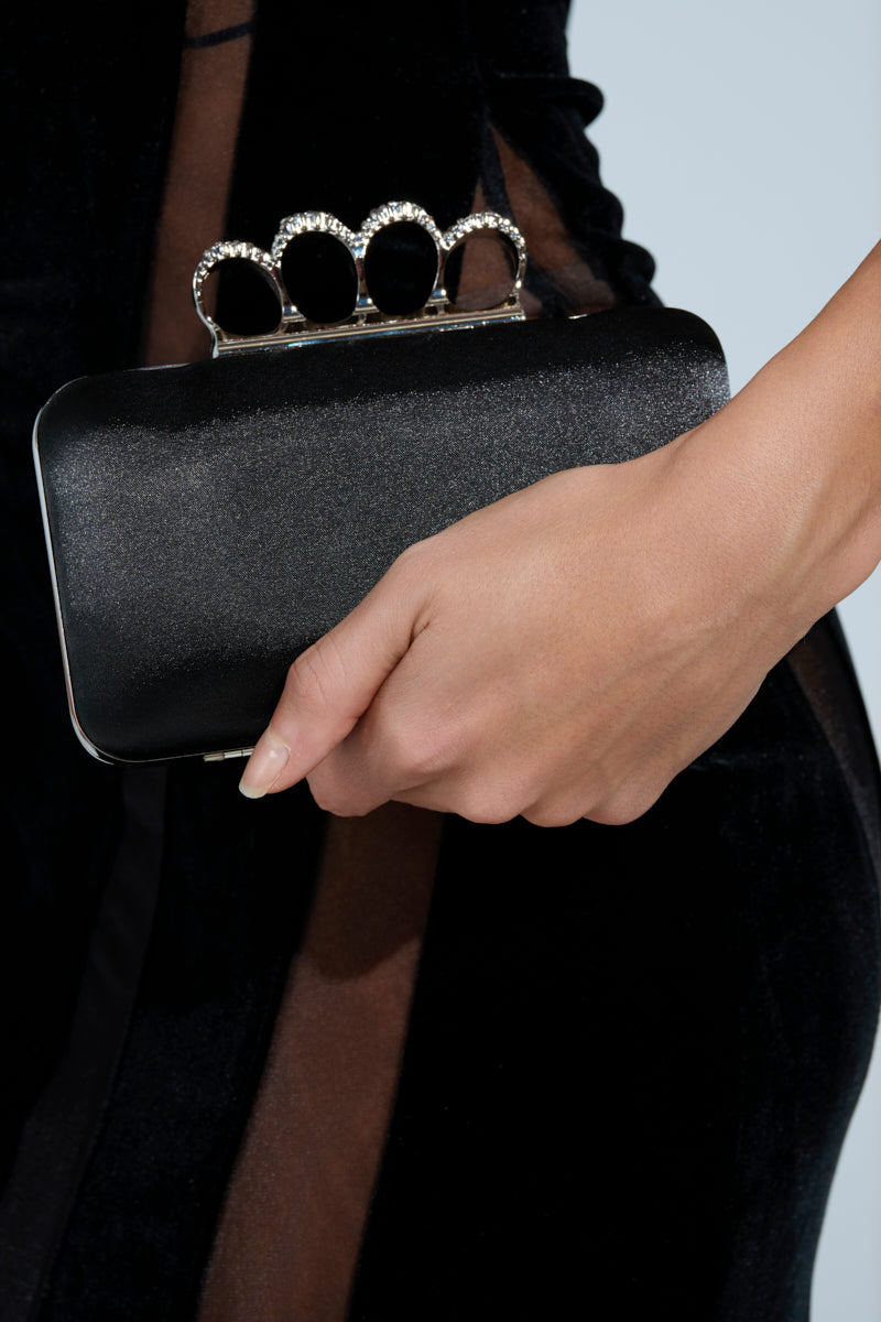 A person holds the RING CLUTCH, a black satin handbag with jeweled rhinestone ring handles, while wearing a black outfit with sheer panels.