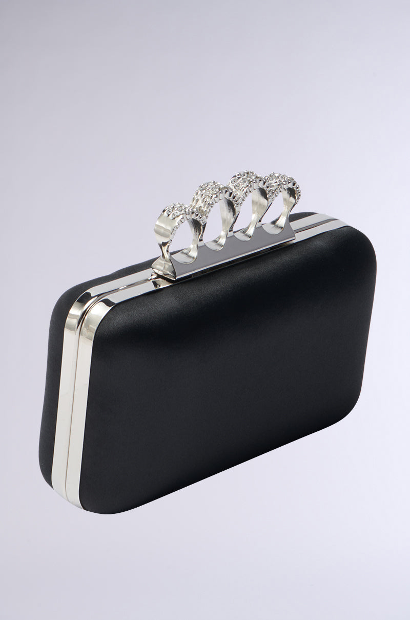 The RING CLUTCH is a black rectangular clutch with silver edges and a decorative handle featuring four ring-like holders encrusted with sparkling rhinestones.
