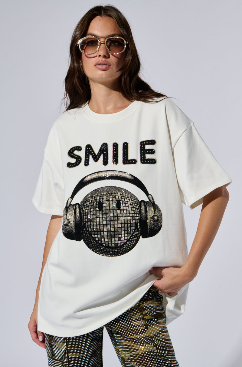 A woman with long hair is wearing large sunglasses and an oversized white graphic tee called the "SMILE RHINESTONE DETAIL OVERSIZED TSHIRT IN WHITE," which features the word "SMILE" in rhinestone detail above a smiling globe wearing headphones. She has one hand in her pocket and is posing against a plain background.
