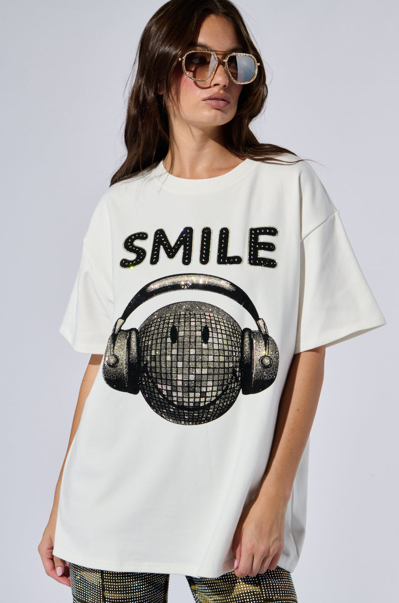 A person with long hair wears sunglasses and the SMILE Rhinestone Detail Oversized T-Shirt in White, featuring the word "SMILE" in rhinestones above a disco ball with headphones. The person stands against a plain background.
