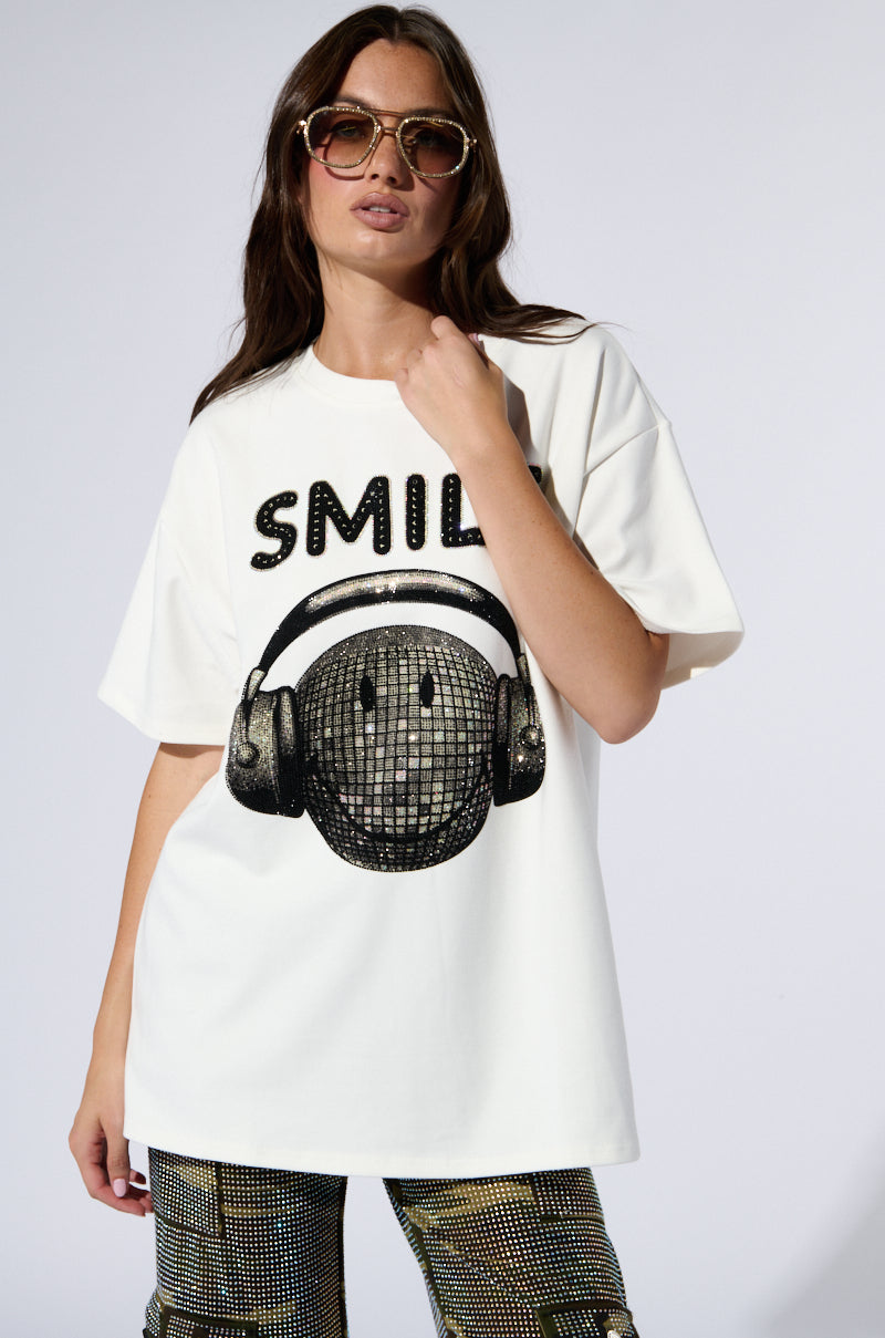 A woman with long brown hair wears oversized sunglasses and a white "SMILE RHINESTONE DETAIL OVERSIZED TSHIRT," which features the word "SMILE" embellished with rhinestones and a smiling face with headphones. She stands confidently against a plain background, one hand resting by her side and the other in her pocket.