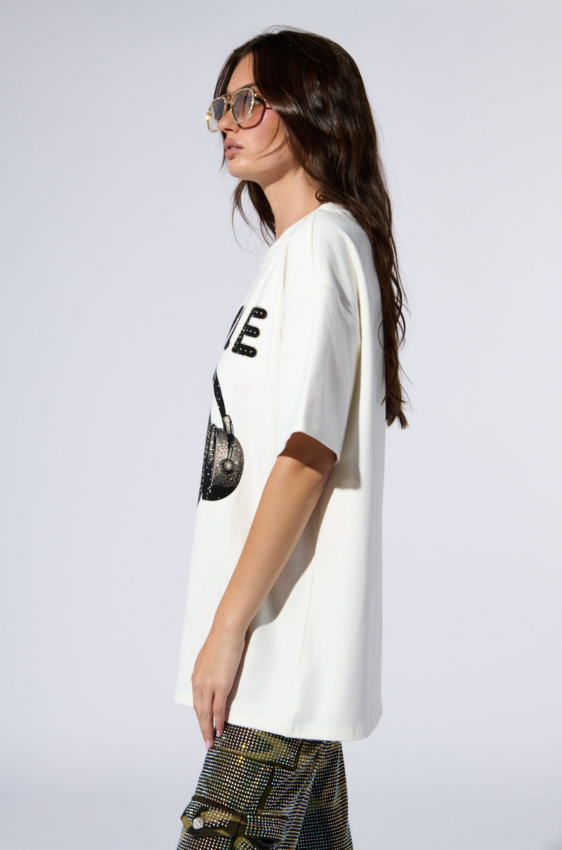 A person with long dark hair wears oversized sunglasses and the SMILE RHINESTONE DETAIL OVERSIZED TSHIRT IN WHITE. They stand in profile against a plain background, with one hand by their side. The white graphic tee features large, bold text, an image of a handbag, and smile rhinestone detail.
