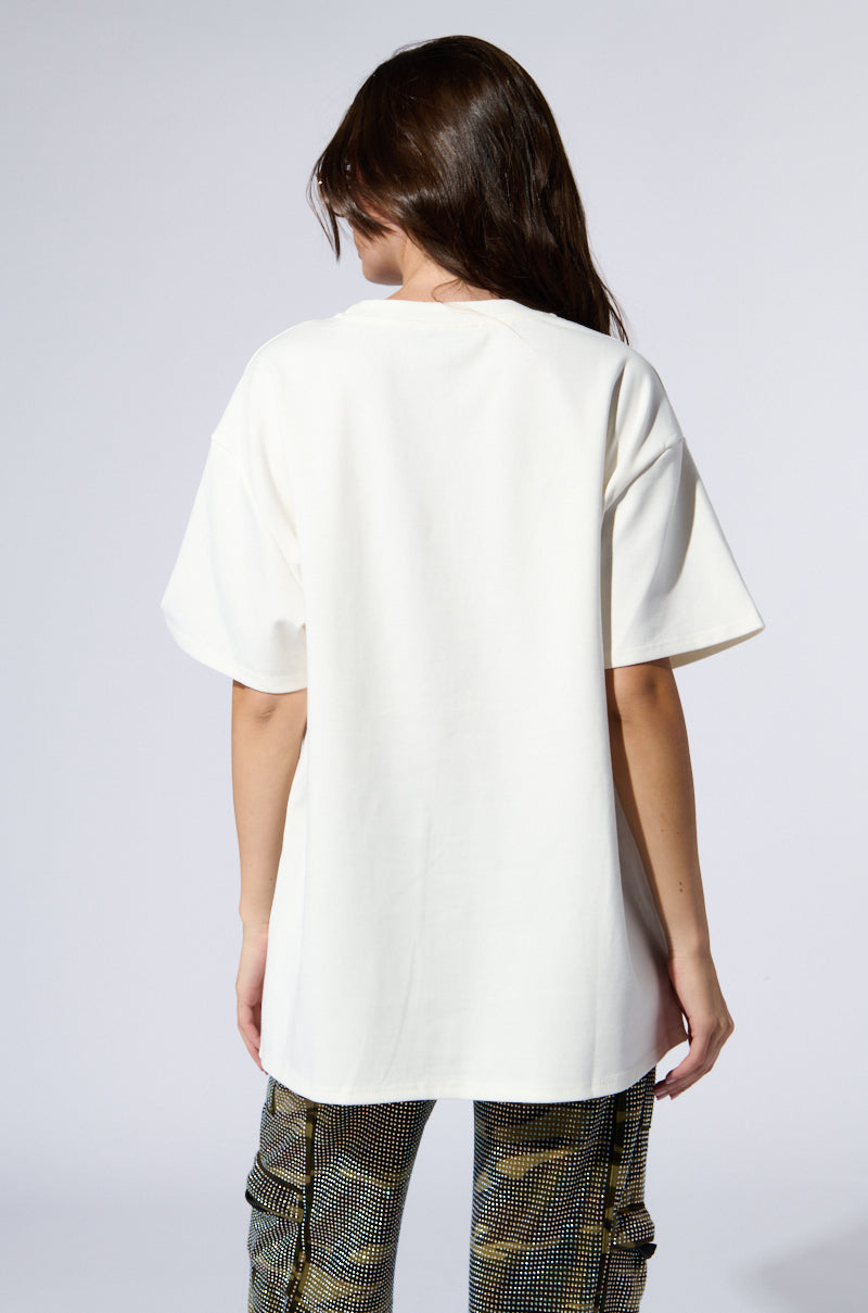 A person with long brown hair is shown from the back, wearing an SMILE RHINESTONE DETAIL OVERSIZED TSHIRT IN WHITE and patterned pants. The background is plain and light-colored.