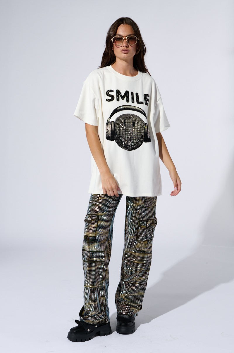A person with long brown hair is wearing sunglasses, the SMILE RHINESTONE DETAIL OVERSIZED TSHIRT IN WHITE featuring a graphic of headphones, and loose, patterned pants with multiple pockets. They stand confidently on a neutral-colored background, hands by their sides.