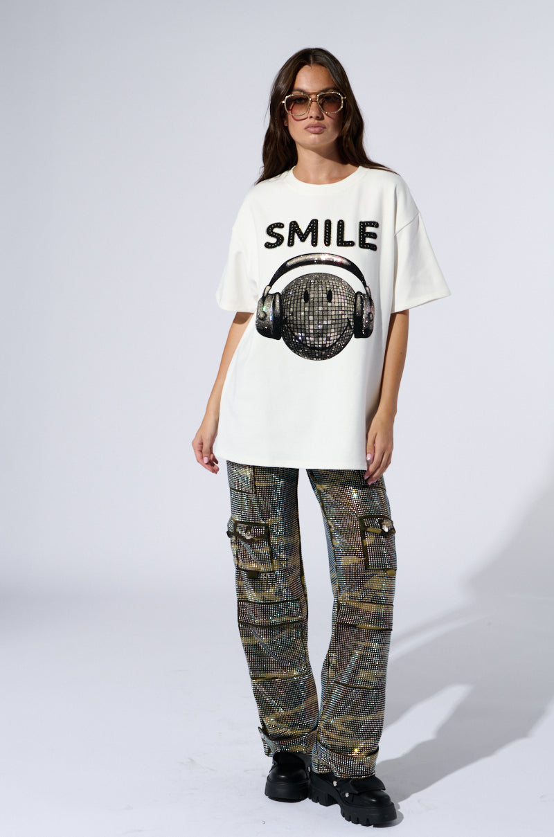 A woman wearing oversized sunglasses and the SMILE RHINESTONE DETAIL OVERSIZED TSHIRT IN WHITE, which features rhinestone details spelling "SMILE" alongside a smiley face with headphones graphic, stands with one hand in her pocket. She pairs the oversized t-shirt with shiny, multicolored cargo pants and black platform shoes against a plain background.