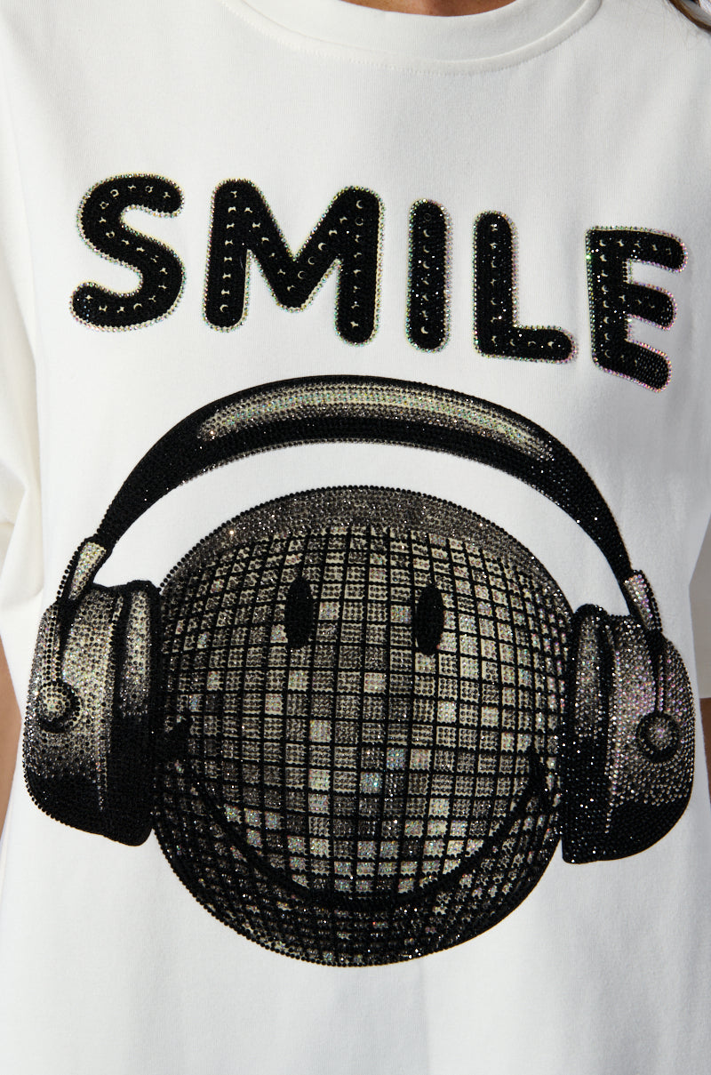 The SMILE Rhinestone Detail Oversized T-Shirt in White features a dazzling graphic of a smiling, spherical face with a grid pattern and large headphones. Above the face, the word "SMILE" is emblazoned in bold, sparkly letters. Ideal for fans of oversized t-shirts with smile rhinestone details.