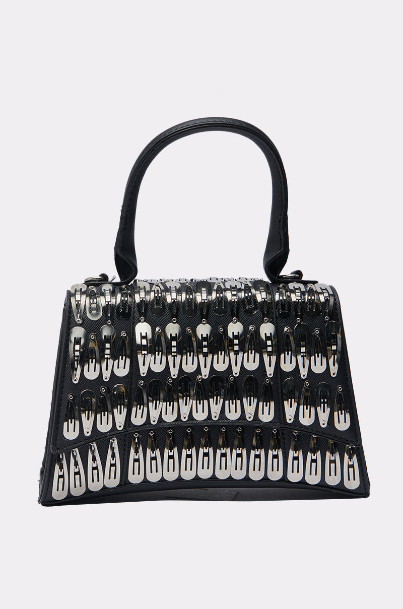 The CLIP IT UP PURSE is a black mini bag with a structured handle, made from faux leather and accented with rows of large silver safety pins on the front and sides for an edgy, bold look.