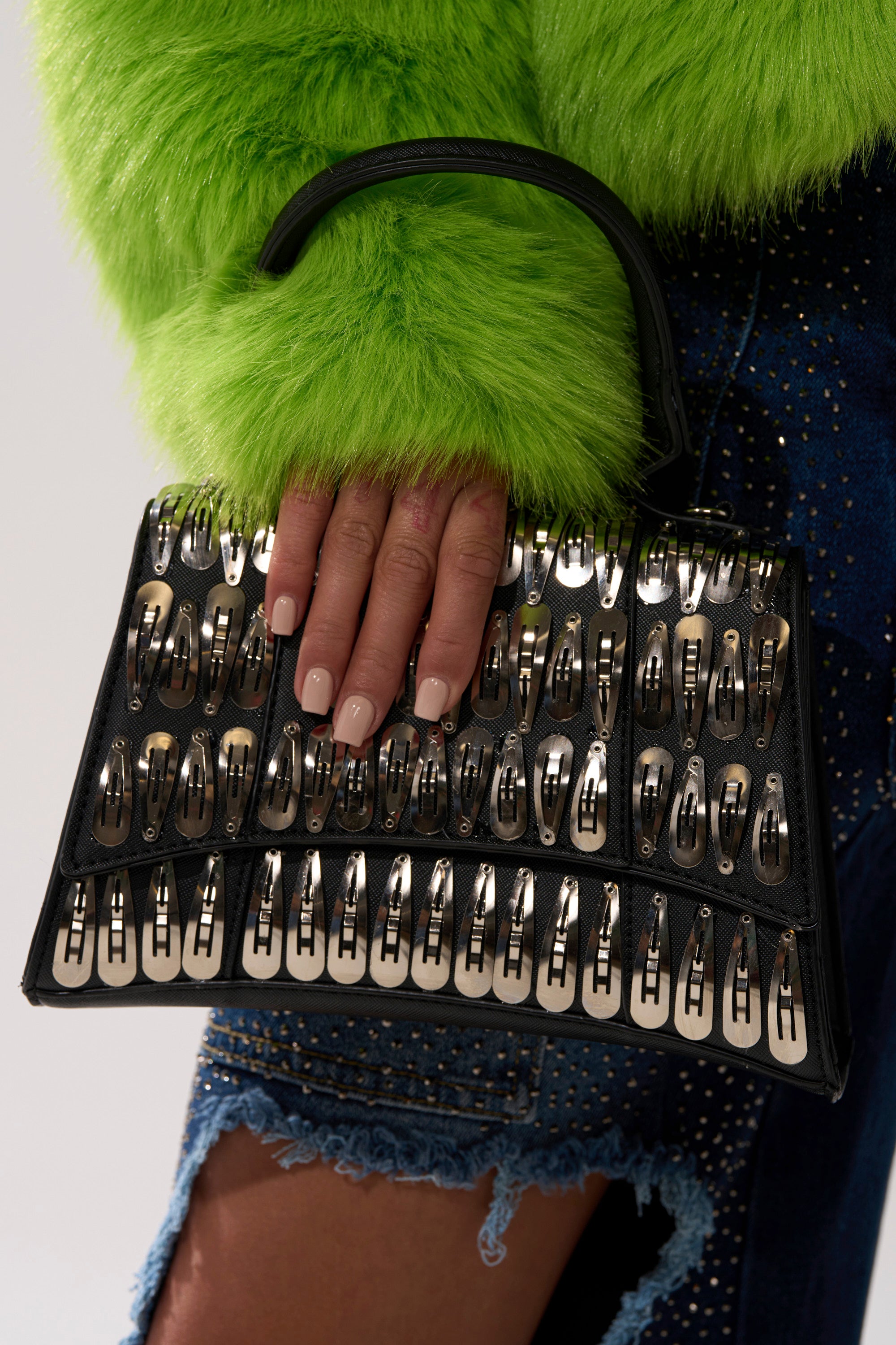 A hand with manicured nails holds the CLIP IT UP PURSE, styled with a bright green faux fur sleeve and frayed blue denim shorts.
