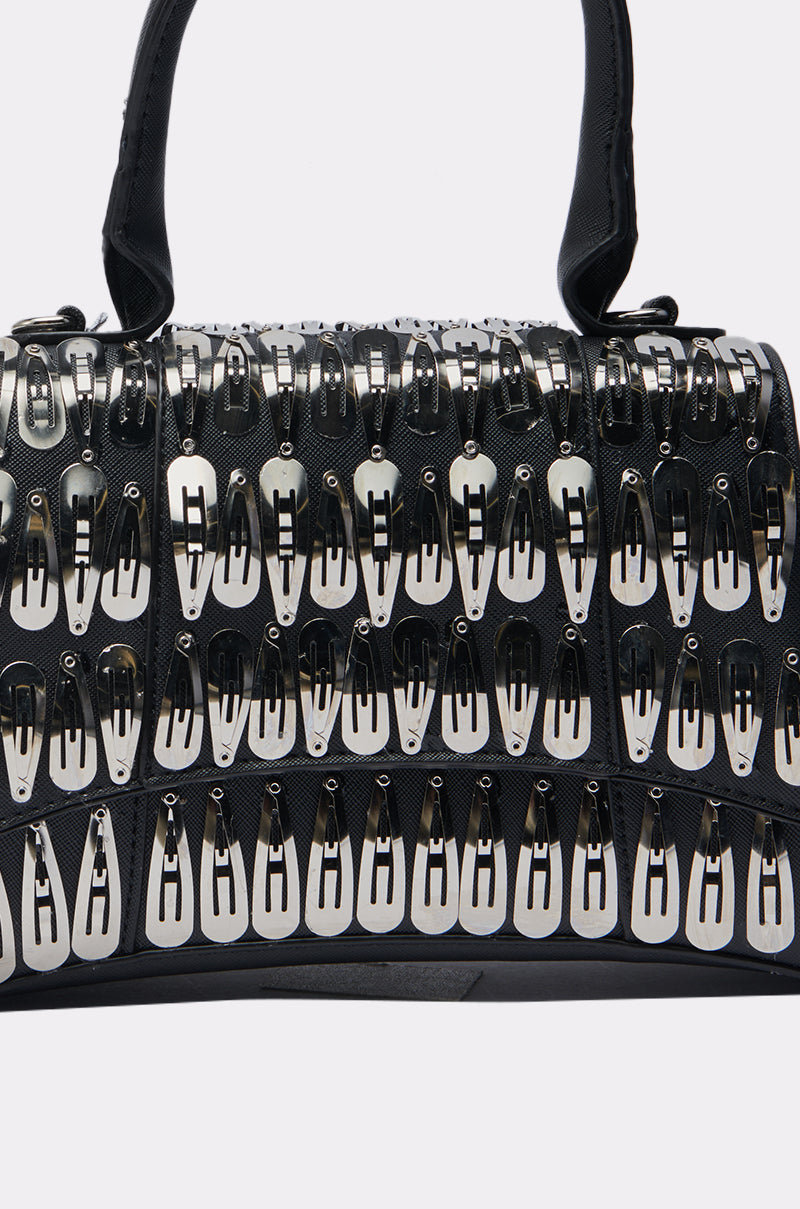 The CLIP IT UP PURSE is a black faux leather mini bag with rows of metallic hair clip embellishments, a structured shape, and two handles for a bold, stylish statement.