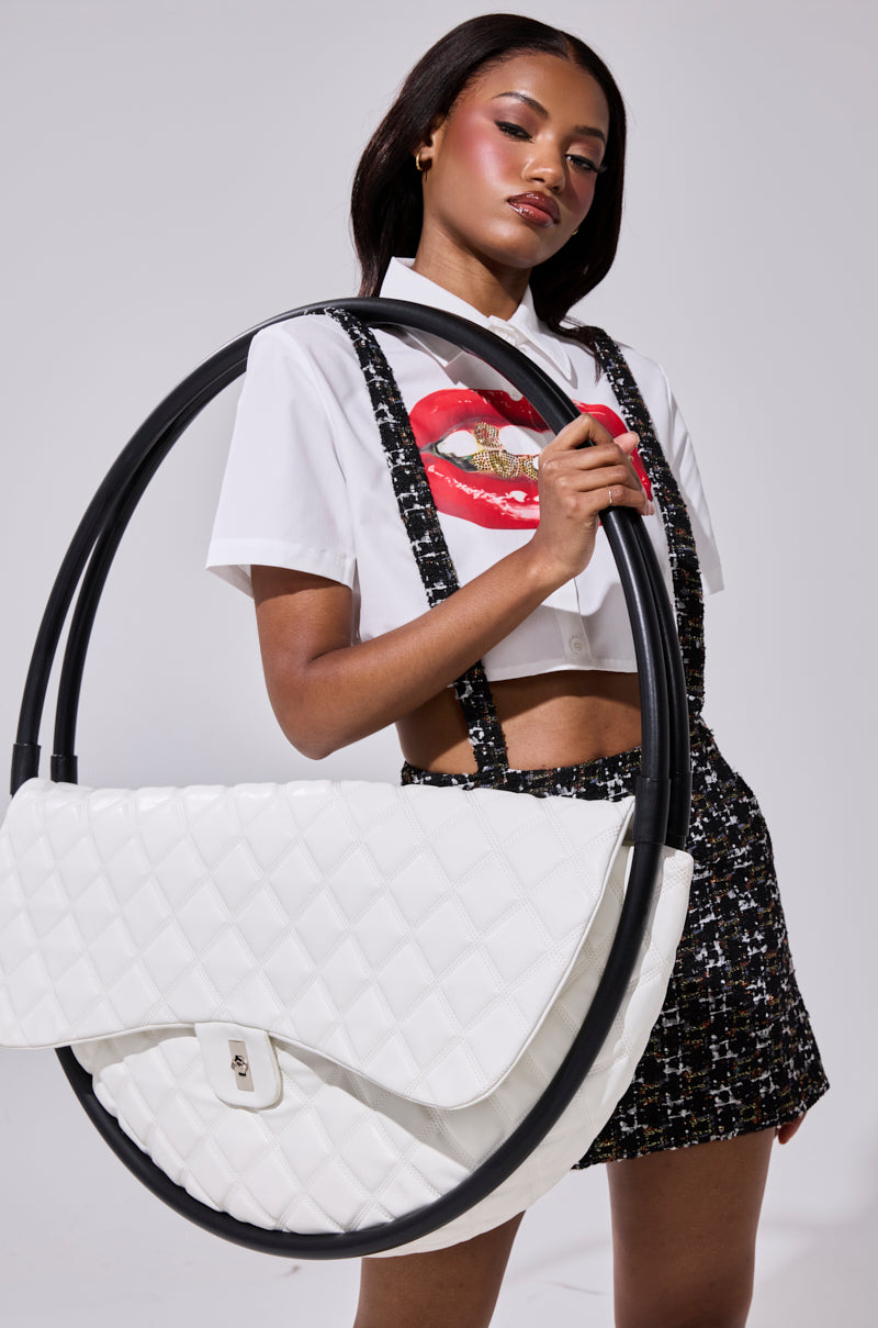 A woman poses confidently holding the IYKYK OVERSIZED PURSE, styled with a white shirt featuring a red lips graphic and a black-and-white tweed suspender skirt.