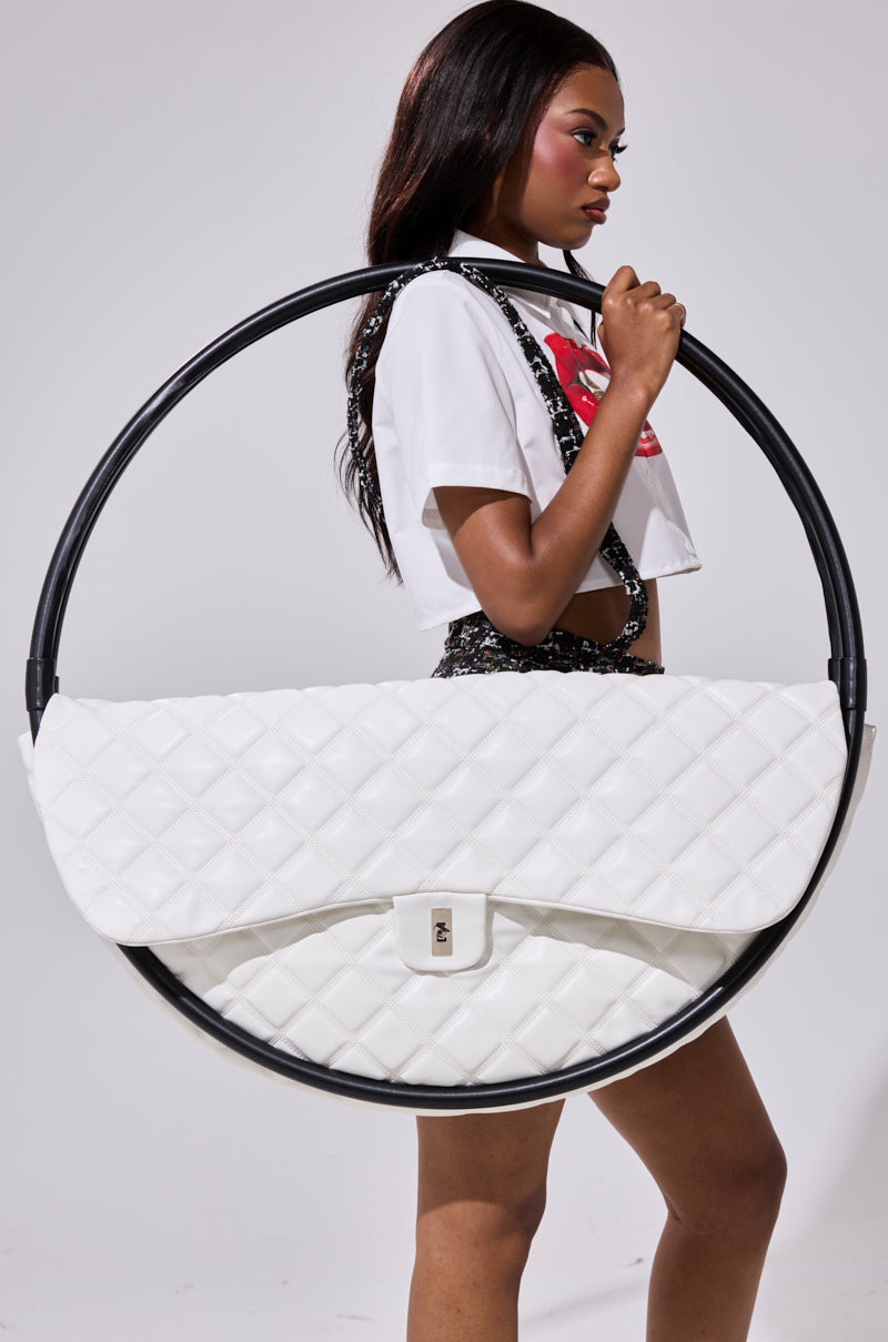 A woman in a white crop top and patterned shorts holds a large circular black frame with the IYKYK OVERSIZED PURSE by AKIRA Label suspended inside, making a bold fashion statement against a plain background.