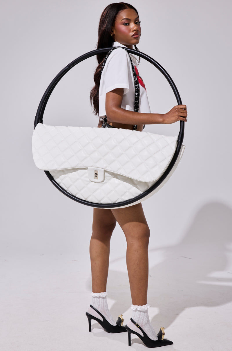 A woman in a white T-shirt, shorts, and black heels stands sideways, holding the IYKYK OVERSIZED PURSE—a circular white quilted faux leather bag with flap, black trim, and strap—against a plain light gray background.