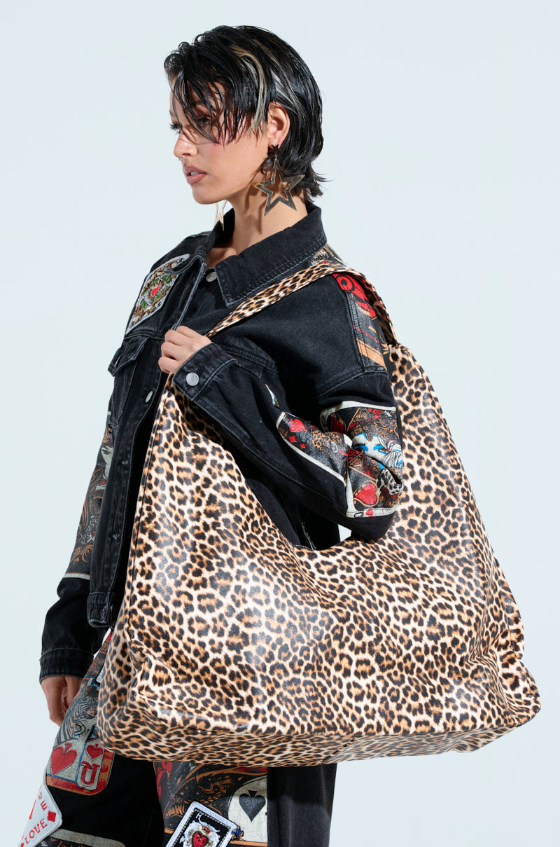 A person with short hair, dressed in a black denim jacket and matching pants adorned with patches, confidently carries the WILD ONE PU OVERSIZED TOTE on their shoulder. The leopard print of the tote stands out strikingly against the stark white background, highlighting their bold style.