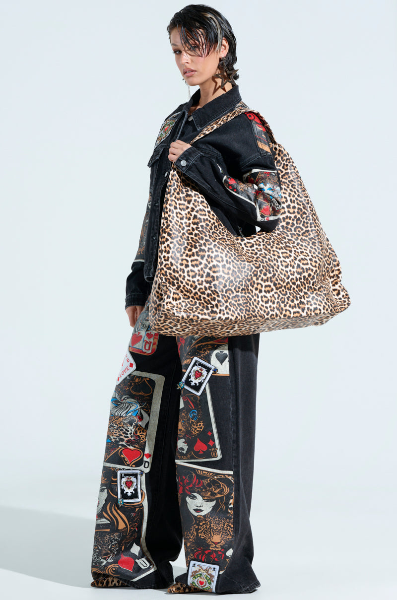 A person in a dark, patterned outfit holds the WILD ONE PU OVERSIZED TOTE with leopard print. Their pants and sleeves display vibrant playing card designs against a plain, light background, accentuating their fashion-forward appearance.