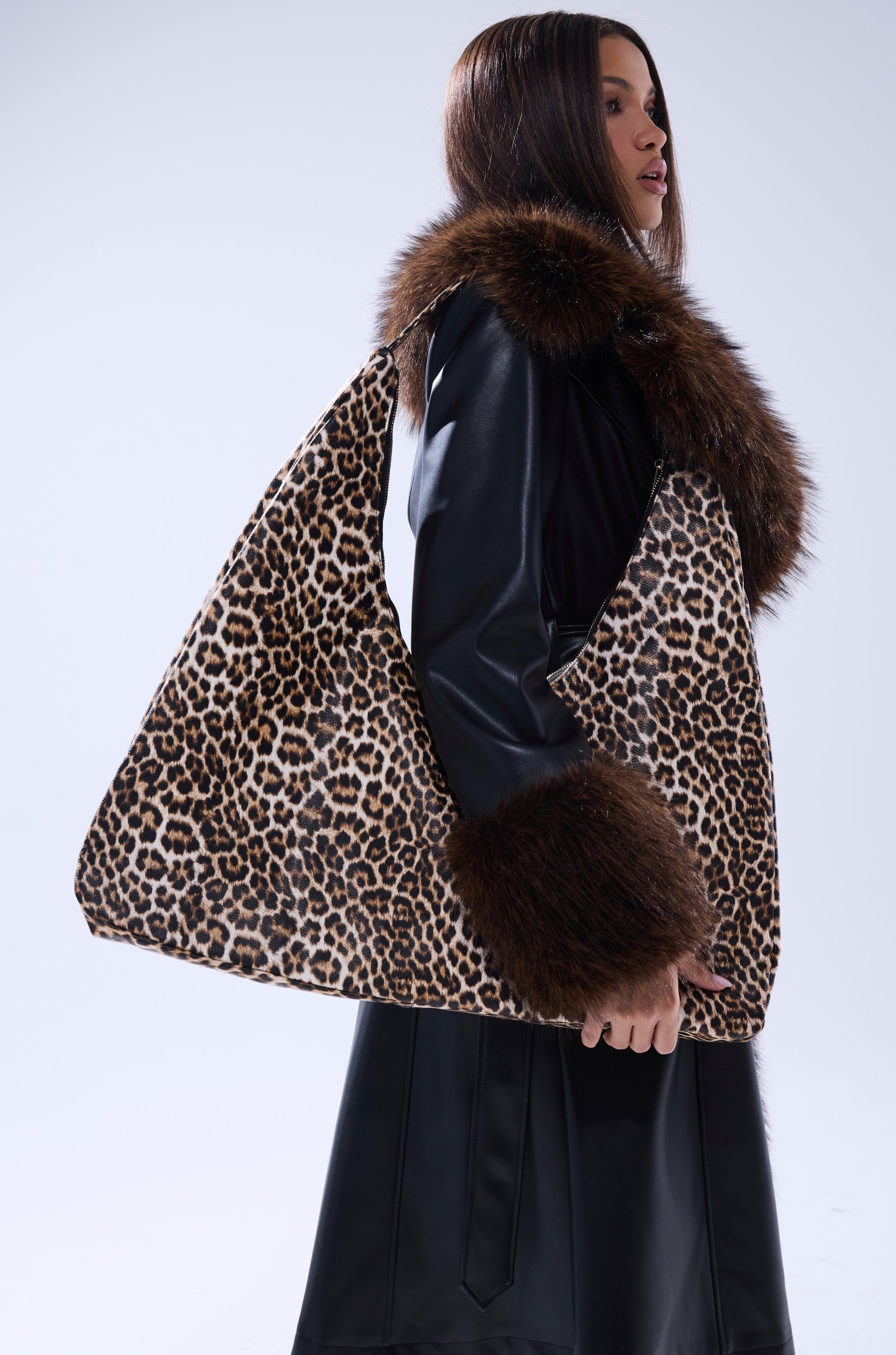 A woman in a black leather coat with brown fur trim stands sideways, holding the WILD ONE PU OVERSIZED TOTE in leopard print against a plain light gray background.