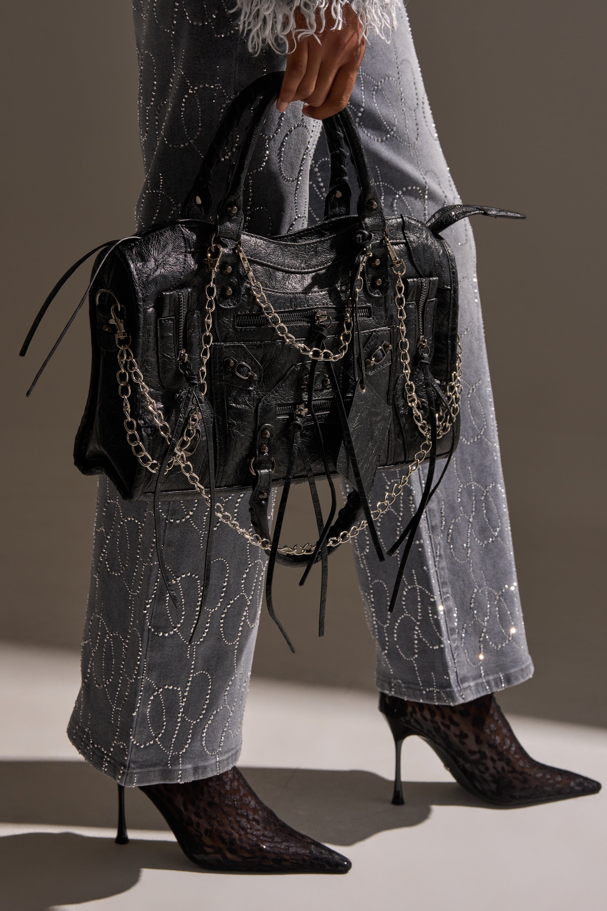 Wearing embellished gray pants and pointed dark heels, a person holds the edgy YOUR LOSS PURSE, featuring silver chains and multiple straps. The neutral background highlights its bold chain details and standout style.