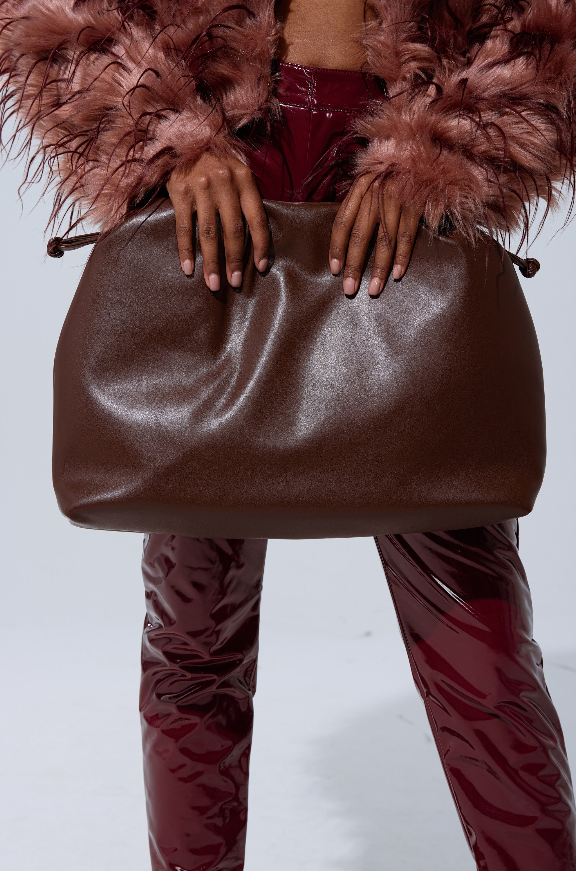 A person in a fluffy pink jacket and shiny burgundy pants holds the IN PARIS WITH IT PURSE, a large, soft faux leather bag, against a plain light background.