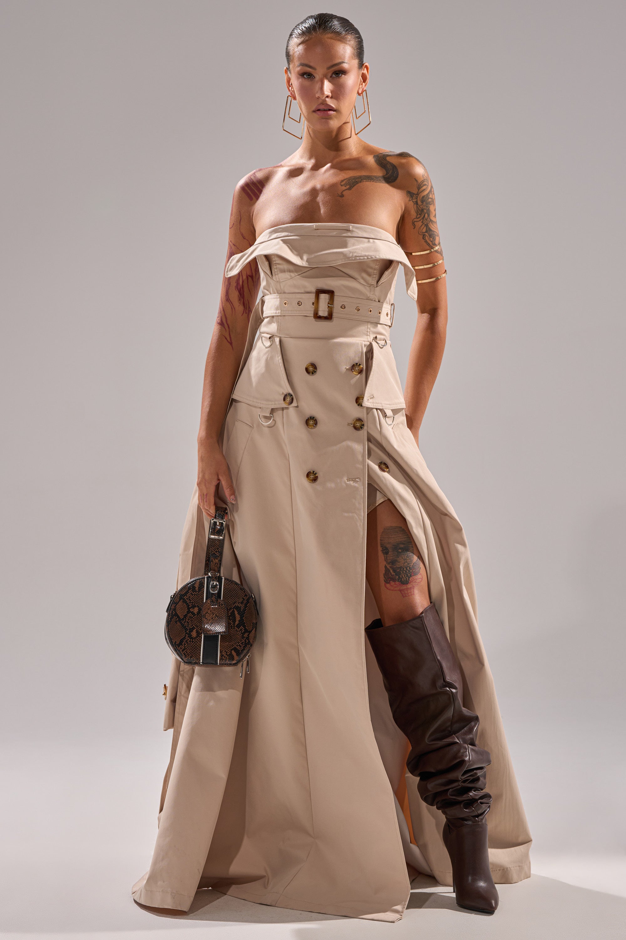A woman exudes confidence in the STRAPLESS TRENCH MAXI DRESS, featuring a belted waist and high slit, paired with brown knee-high boots, a small round handbag, bold earrings, tattoos, and sleek hair.