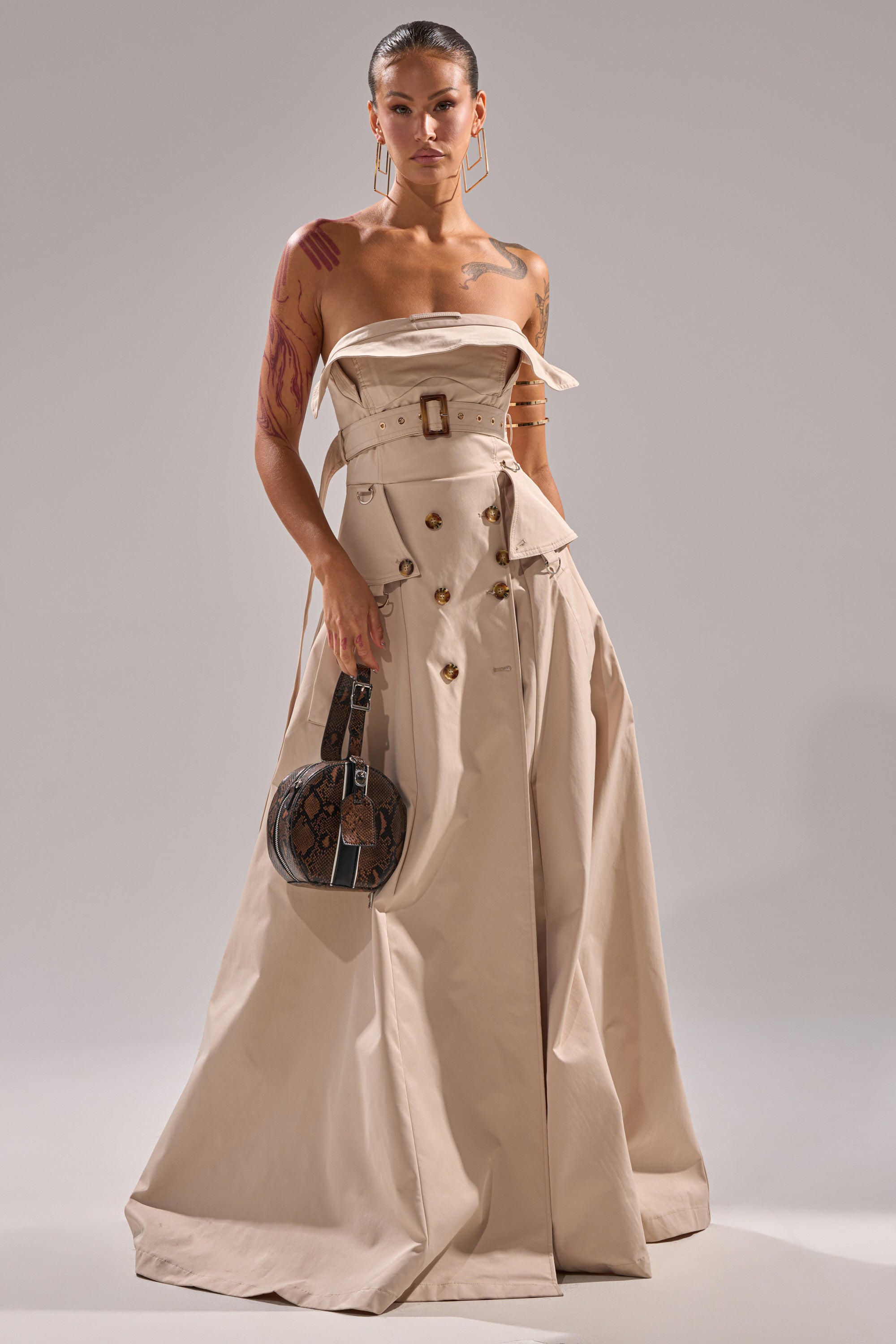 A woman wears the STRAPLESS TRENCH MAXI DRESS in beige, featuring button and belt details, styled like a trench coat. She carries a round, brown patterned handbag and displays arm tattoos, set against a plain light gray background.