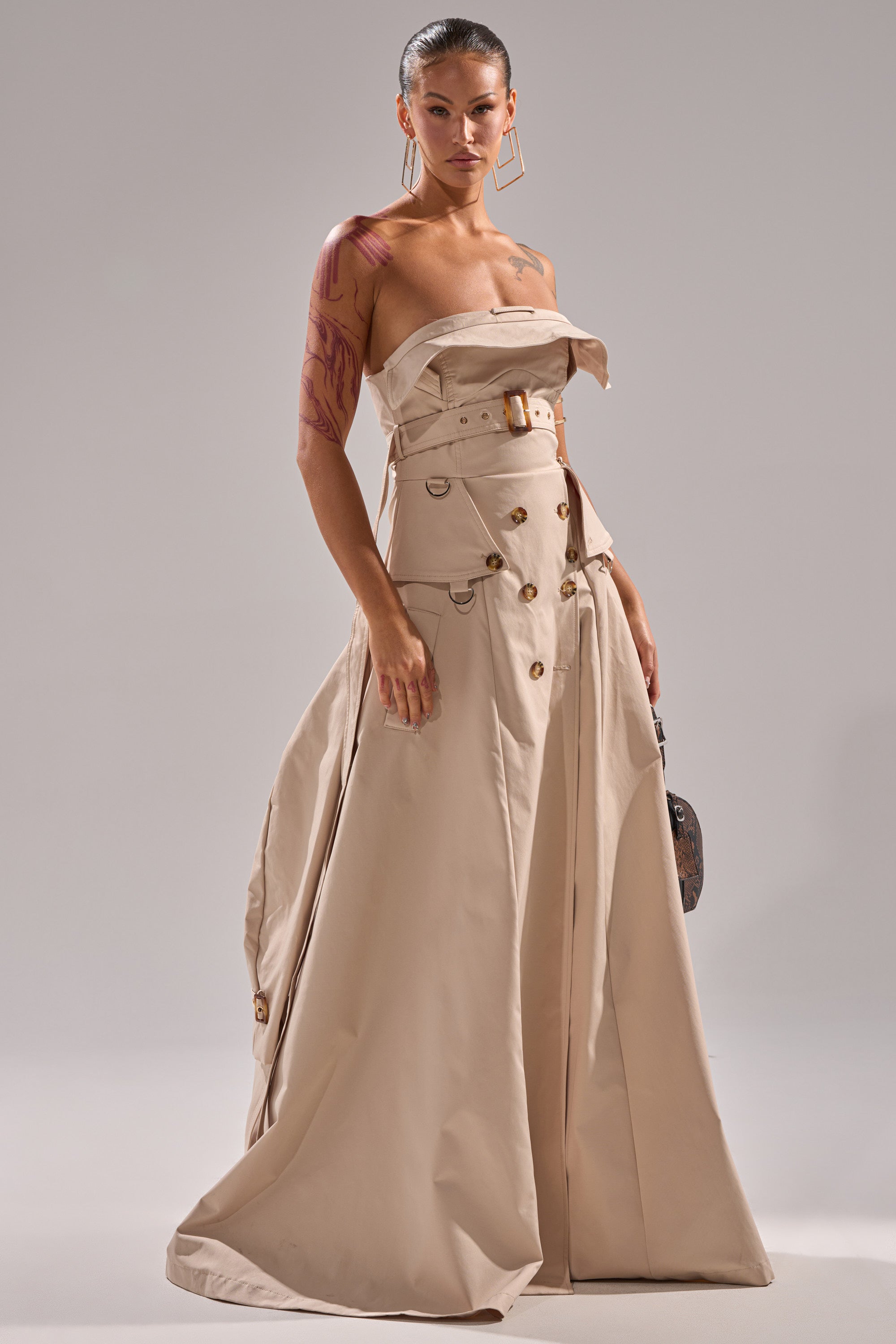 A woman models the STRAPLESS TRENCH MAXI DRESS in a studio, featuring buttons, a belted waist, and large lapels. She accessorizes with geometric earrings, has slicked-back hair, and a visible tattoo on her left arm.