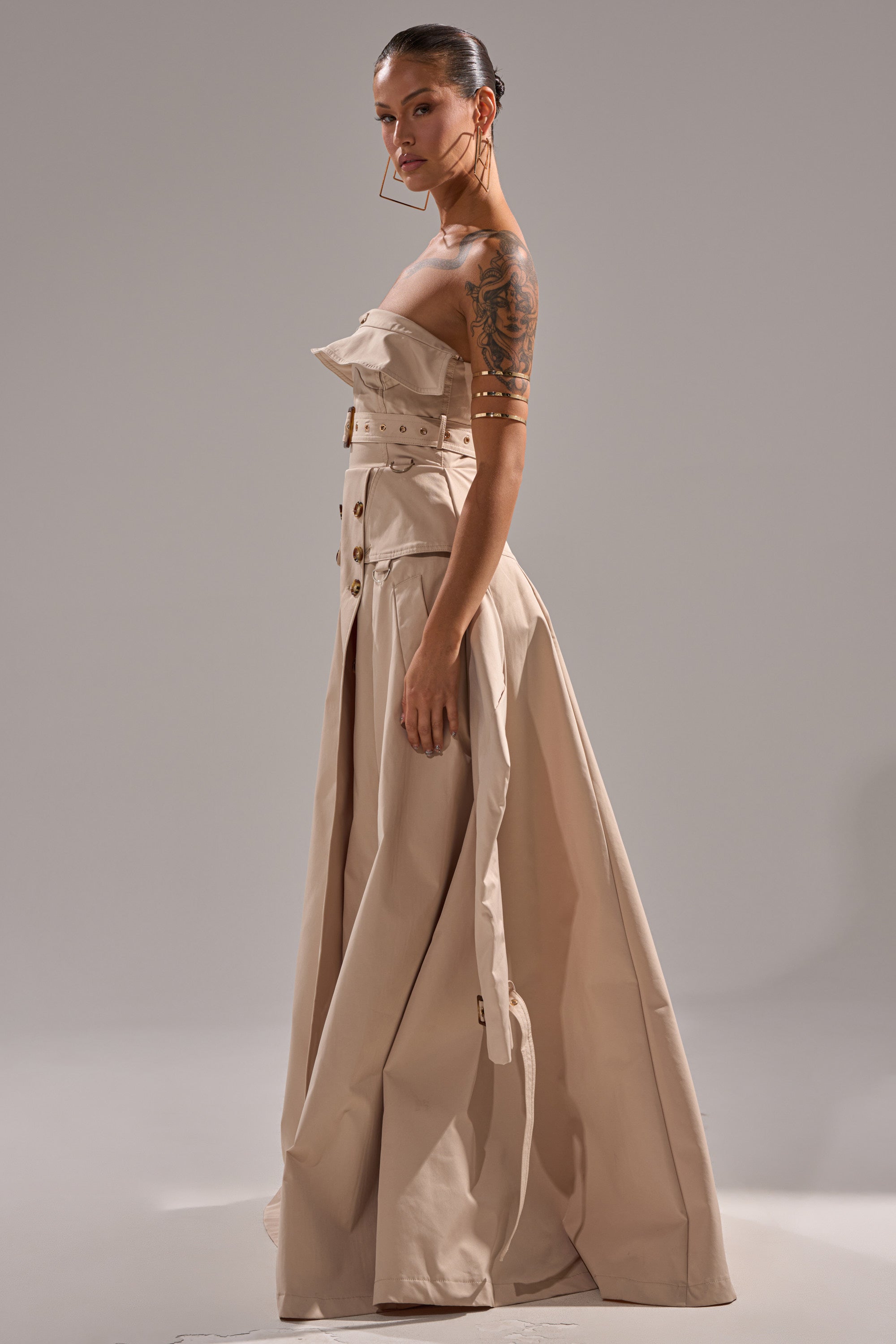 A woman stands sideways in the STRAPLESS TRENCH MAXI DRESS, featuring a beige hue, structured bodice, decorative buttons, and strapless design. Her slicked-back hair shows off an arm tattoo and large hoop earrings.