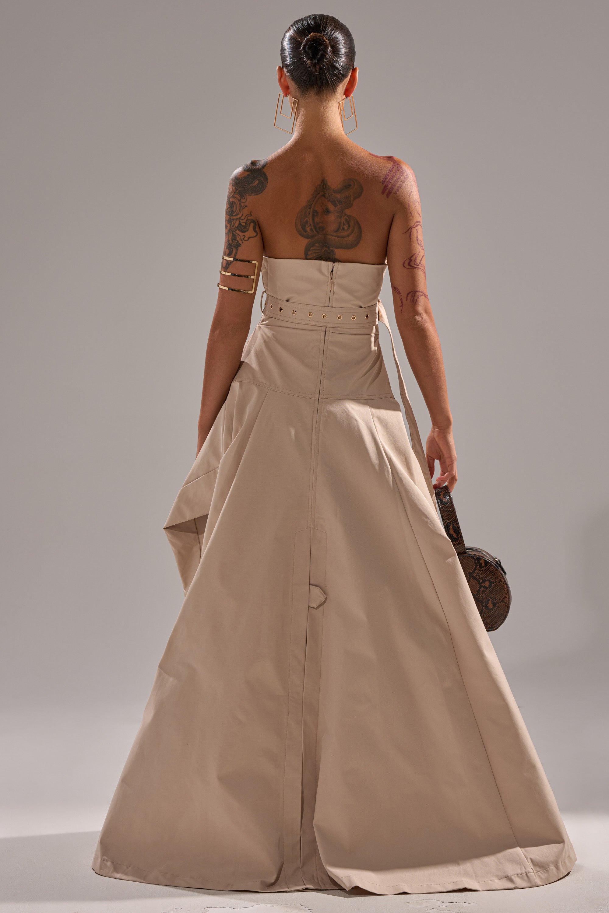 A tattooed woman, seen from behind in a STRAPLESS TRENCH MAXI DRESS with a flared skirt, holds a round brown bag. Her hair is styled in a sleek bun against a plain neutral background.