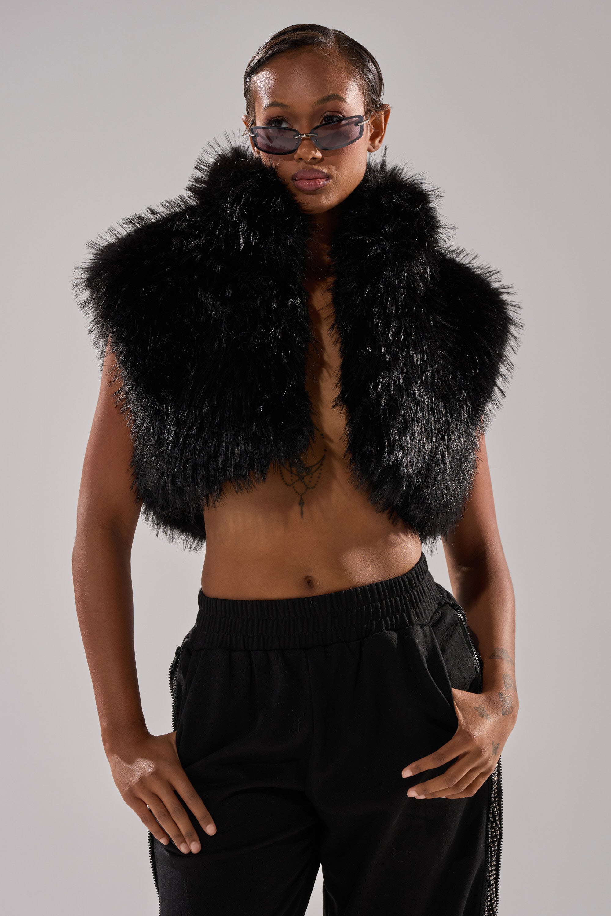 Wearing the COCO CROPPED FUR VEST IN BLACK, dark sunglasses, and black pants, a person with short, slicked-back hair stands confidently against a plain background with their hands in their pockets.