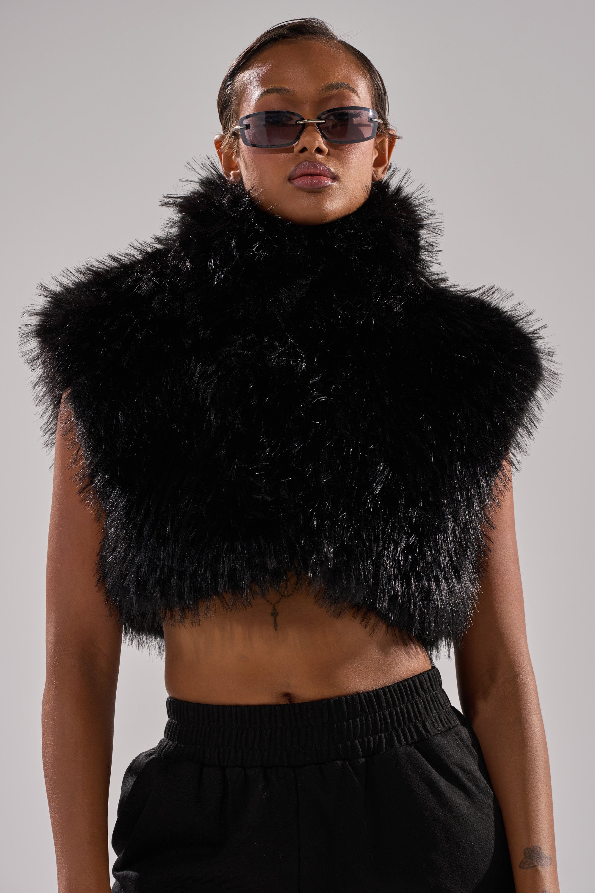 A person models the COCO CROPPED FUR VEST IN BLACK with sunglasses and black pants, standing against a plain background and looking confidently at the camera.