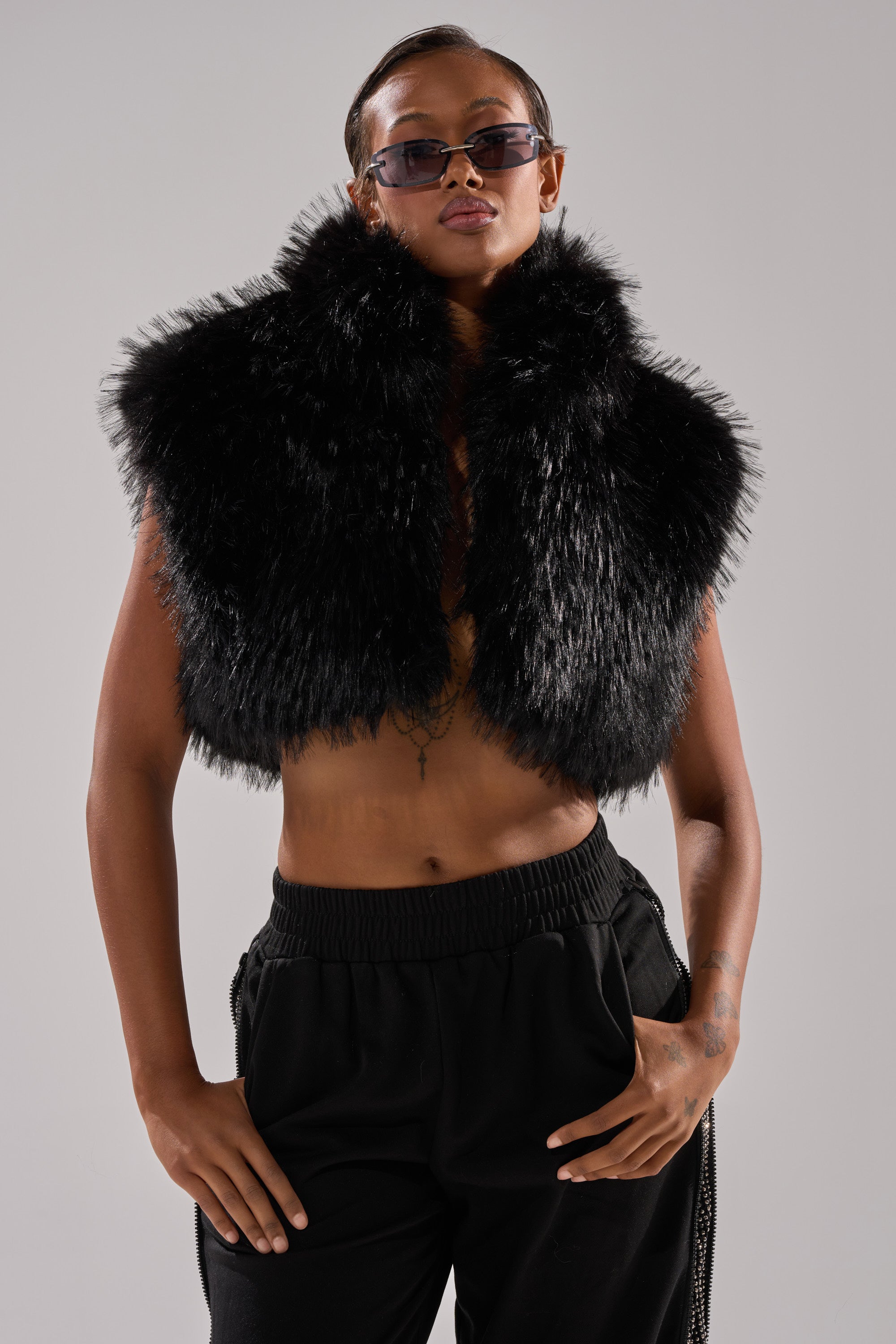 A person confidently poses against a plain light background, wearing the COCO CROPPED FUR VEST IN BLACK, styled without a shirt and paired with black pants and sunglasses.