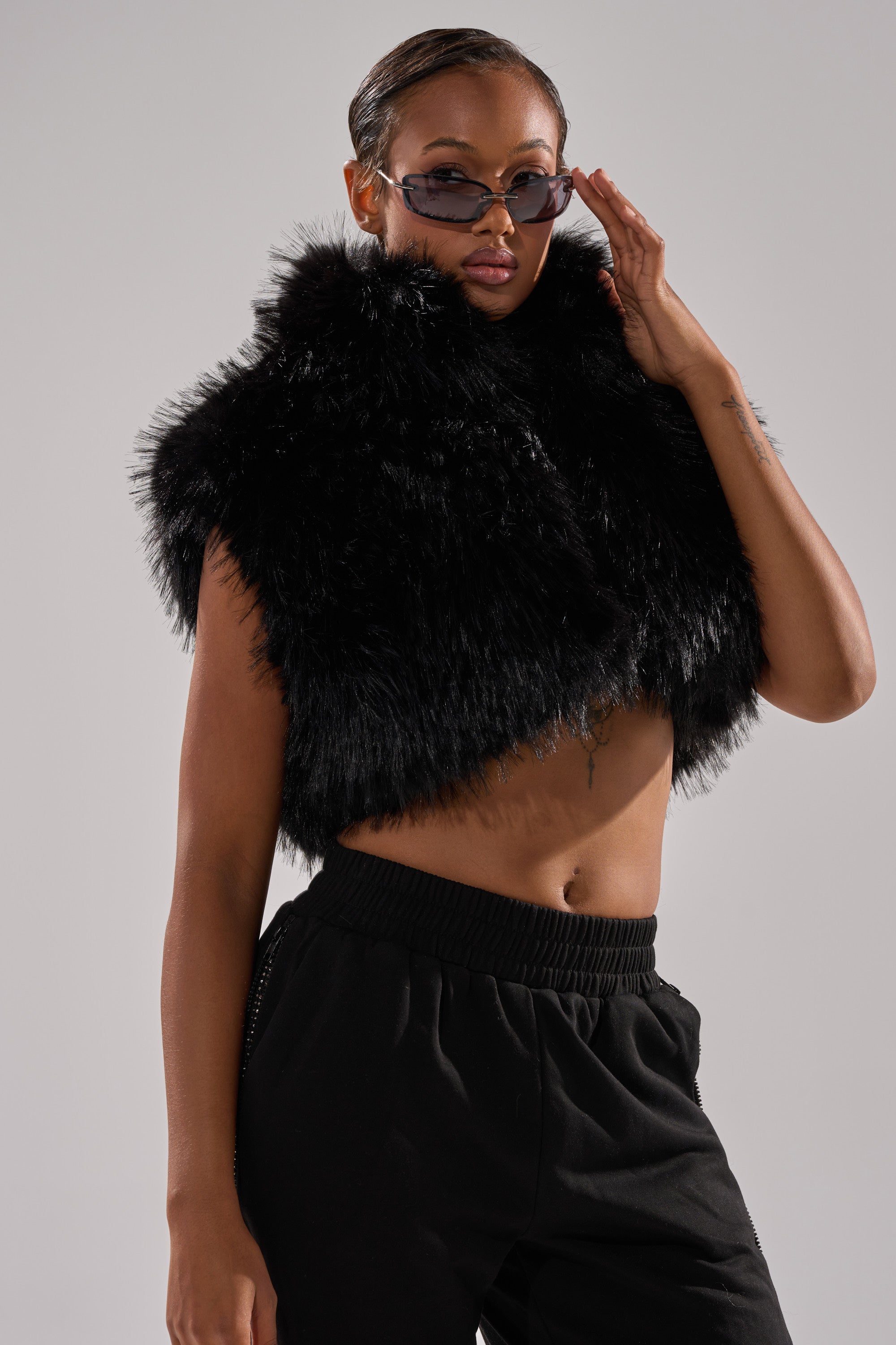 A woman poses confidently against a plain light gray background, wearing dark sunglasses, black pants, and the COCO CROPPED FUR VEST IN BLACK.