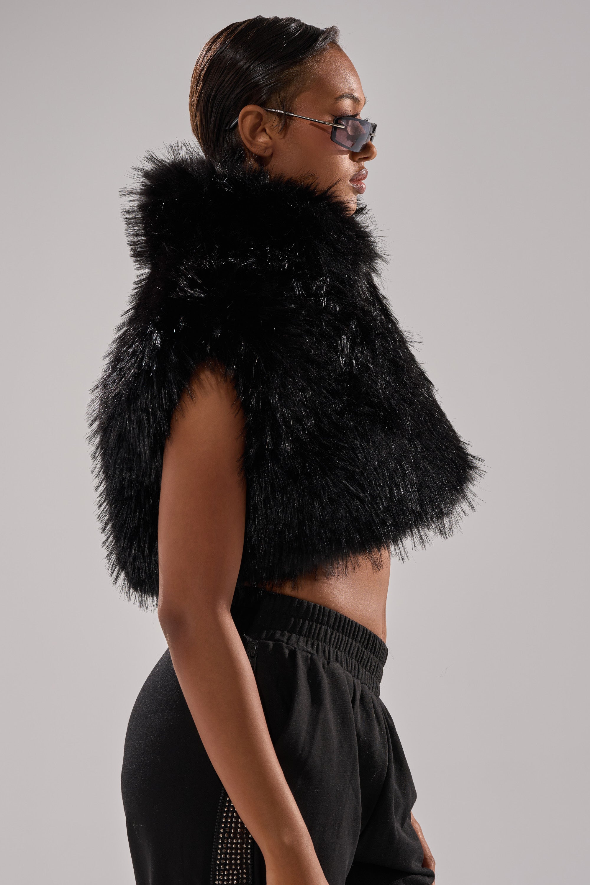 A woman in black sunglasses models the COCO CROPPED FUR VEST IN BLACK with black pants, standing in profile against a light gray background.