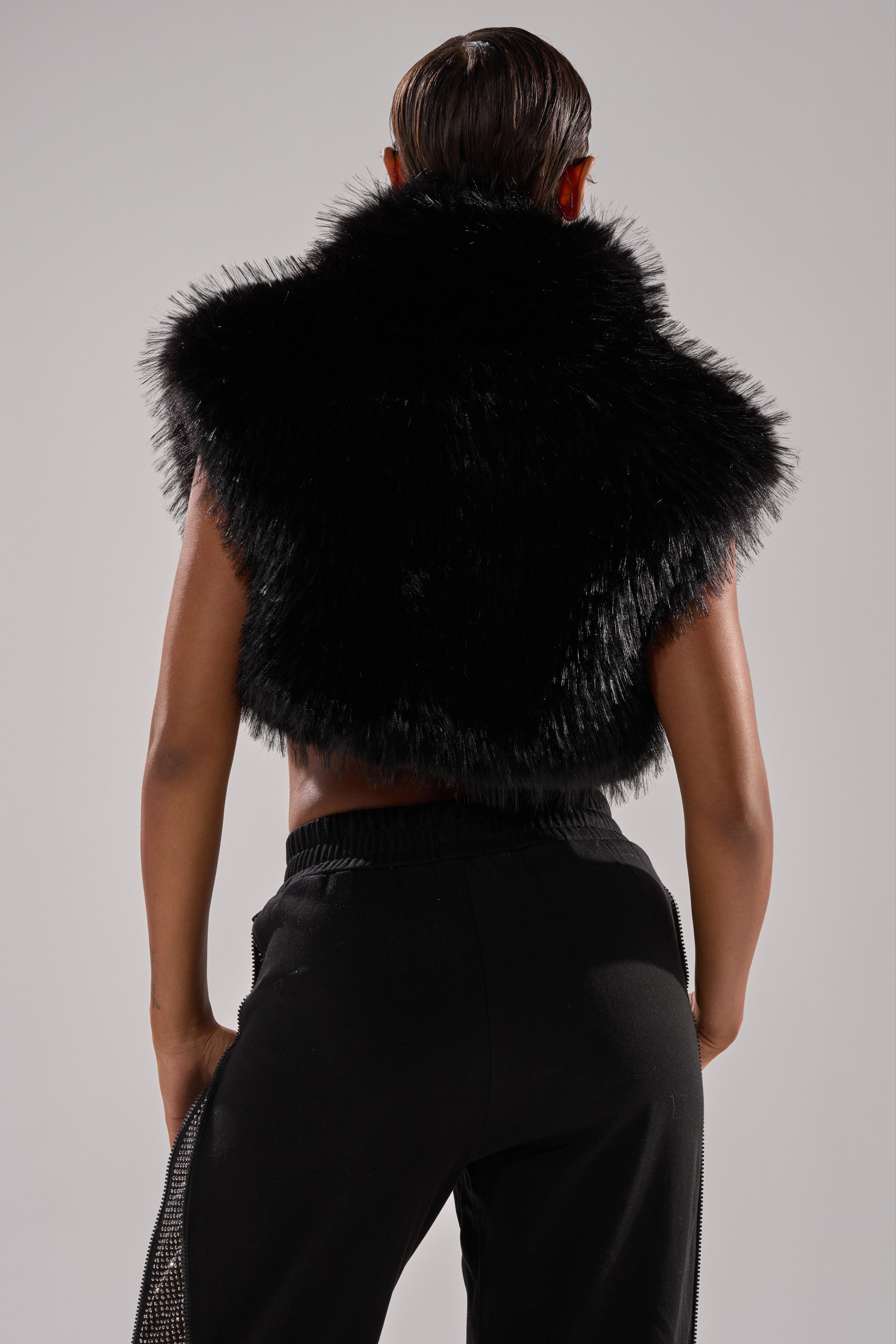 A person faces away from the camera, wearing the COCO CROPPED FUR VEST IN BLACK and black pants, set against a plain light gray background.
