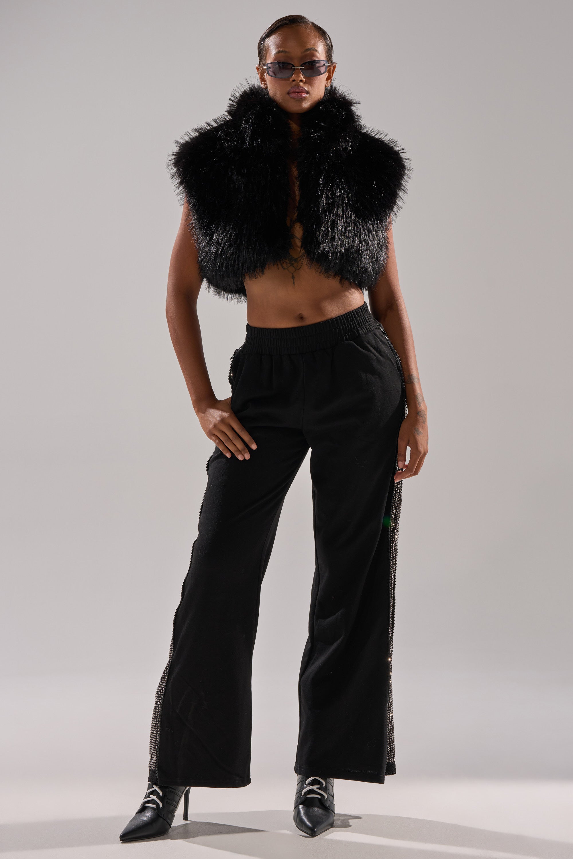 A woman stands confidently in the COCO CROPPED FUR VEST IN BLACK, paired with dark sunglasses, black wide-leg pants, and pointed-toe heels, posing with one hand on her hip against a light gray background.