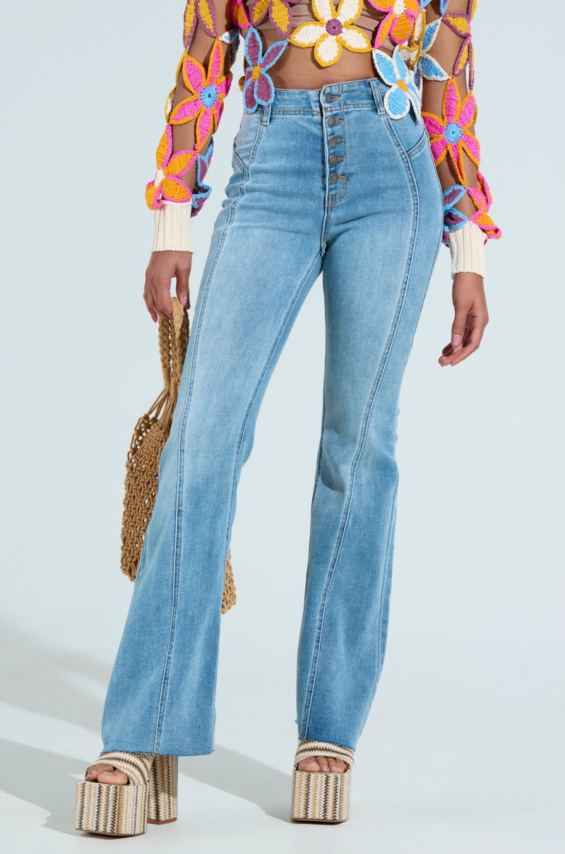 Wearing SOPHIA FLARE LEG DENIM JEANS, a person in high-waisted, button-fly light-wash denim with a slight flare flaunts a colorful floral crochet top. They hold a woven bag and wear platform sandals with striped soles against a plain light blue background.