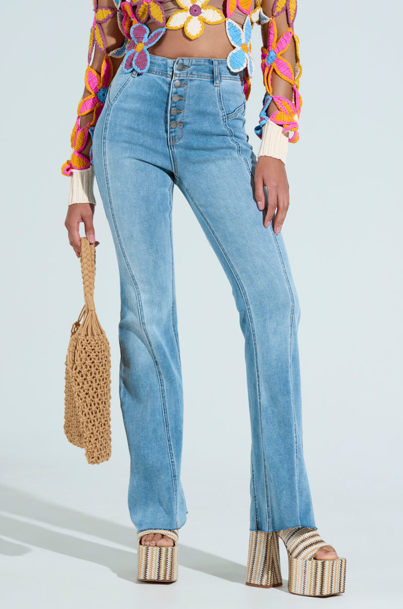A person wearing SOPHIA FLARE LEG DENIM JEANS, a colorful floral crochet top, and platform striped heels holds a woven beige handbag against a plain light background.