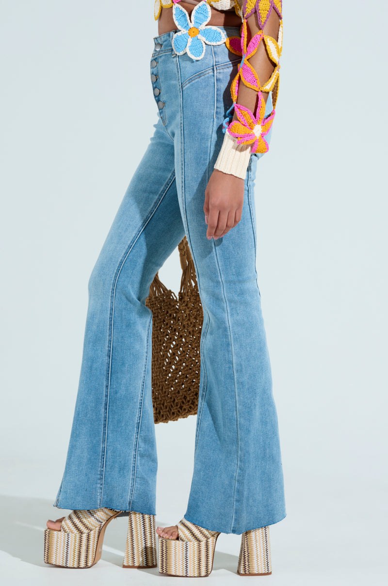 Wearing SOPHIA FLARE LEG DENIM JEANS, someone pairs them with a vibrant floral crochet top and high platform sandals. They accessorize with a woven bag against a simple background, displaying effortless style.