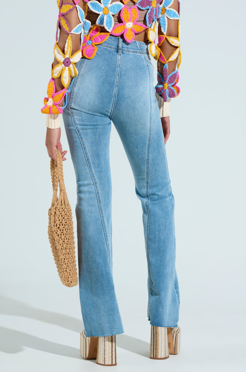 A person is seen from the back wearing SOPHIA FLARE LEG DENIM JEANS in light blue. They sport a colorful floral crochet top, paired with platform heels and a woven bag, set against a light, neutral background.