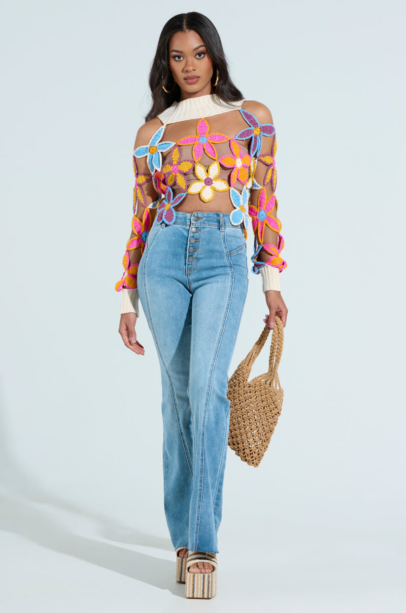 In SOPHIA FLARE LEG DENIM JEANS and platform sandals, a woman carries a woven bag. Her colorful top has large multicolored flowers with a sheer design. Standing against a plain, light background, she has long wavy hair.