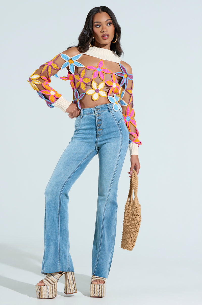 A woman models a colorful floral-patterned mesh sweater with SOPHIA FLARE LEG DENIM JEANS and platform sandals, holding a crocheted handbag against a light blue background.