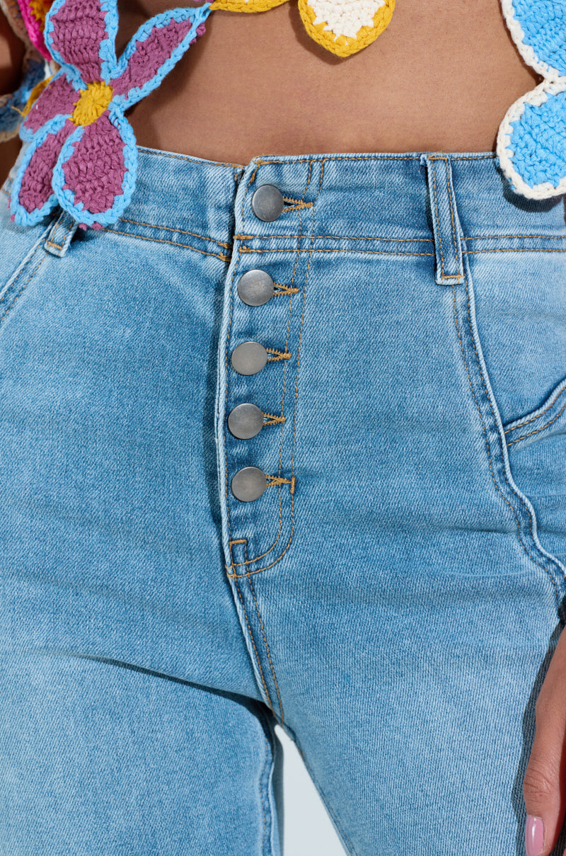Close-up of a person wearing SOPHIA Flare Leg Denim Jeans, featuring a light blue, high-rise fit with five metallic buttons. The top displays crocheted flowers in purple, yellow, and blue against a neutral backdrop.