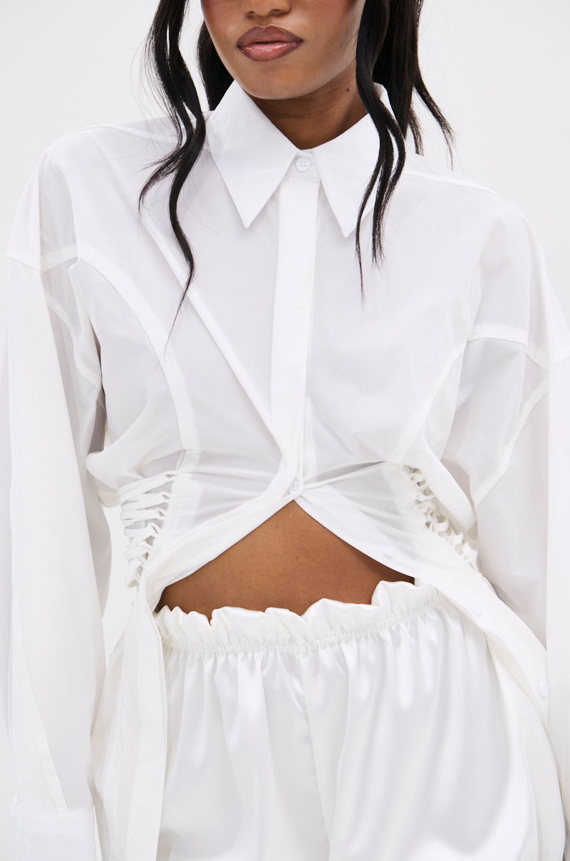 A woman wears the SHAR BUTTON DOWN WITH SIDE TIES, a white blouse with ruched sides and an asymmetrical midriff-baring hem, styled with loose white high-waisted pants for a chic, modern look.
