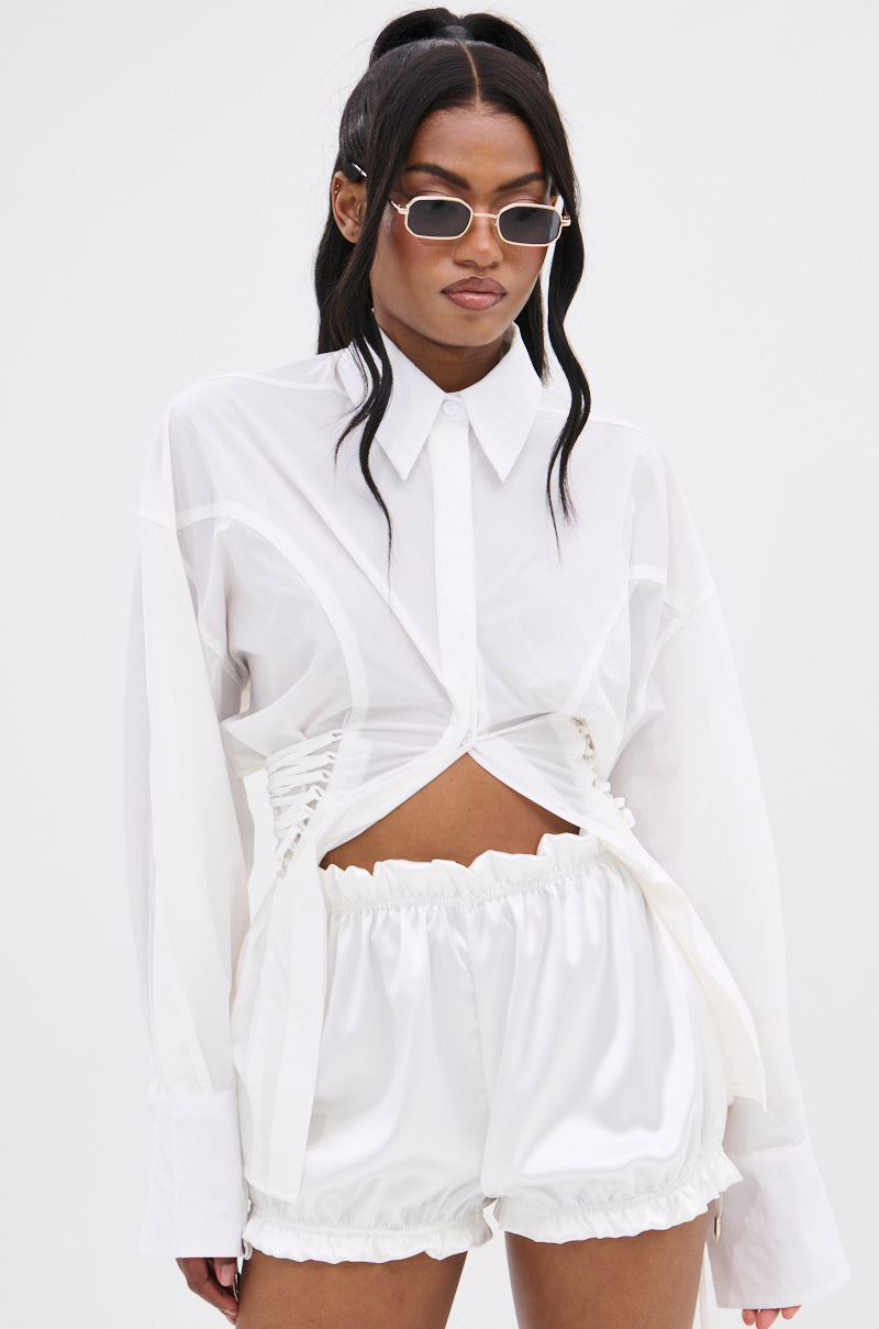 A woman wearing narrow sunglasses, the SHAR BUTTON DOWN WITH SIDE TIES featuring ruched cut-out details for a corset-inspired look, and shiny white loose shorts stands against a plain white background.