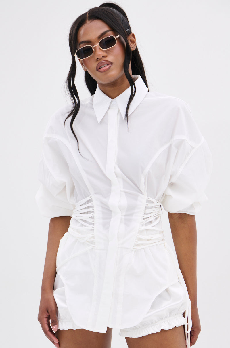 A woman wearing the SHAR BUTTON DOWN WITH SIDE TIES, featuring white fabric, puffed sleeves, and a corset-inspired fit. She accessorizes with slim sunglasses and has long wavy dark hair, standing against a plain white background.