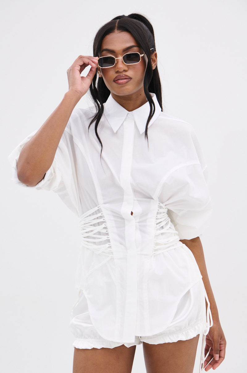 A woman wears the SHAR BUTTON DOWN WITH SIDE TIES—a sheer white blouse with a corset-inspired silhouette and matching shorts—paired with small rectangular sunglasses, standing against a plain white background.