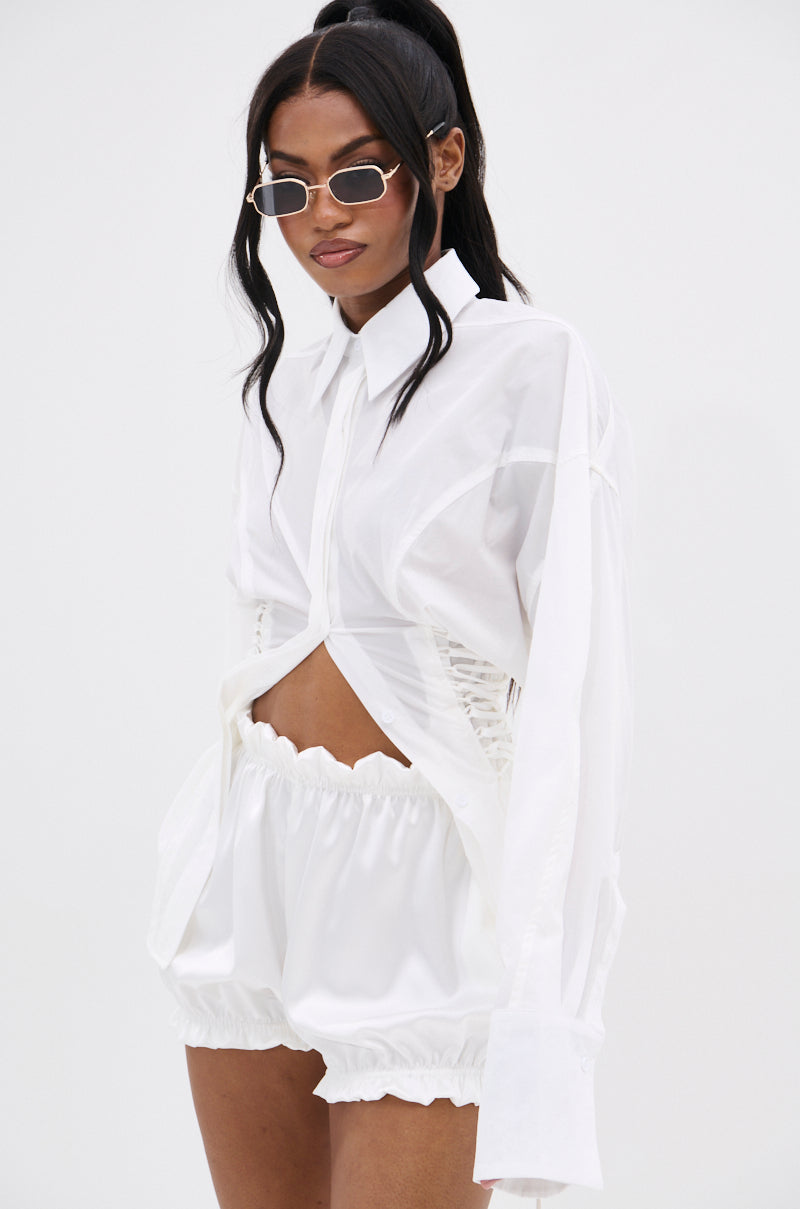 A woman wears the AKIRA Label SHAR BUTTON DOWN WITH SIDE TIES, a sheer white long-sleeve shirt with ruched sides, paired with high-waisted ruffled shorts. She has long dark hair, sunglasses, and a confident look.