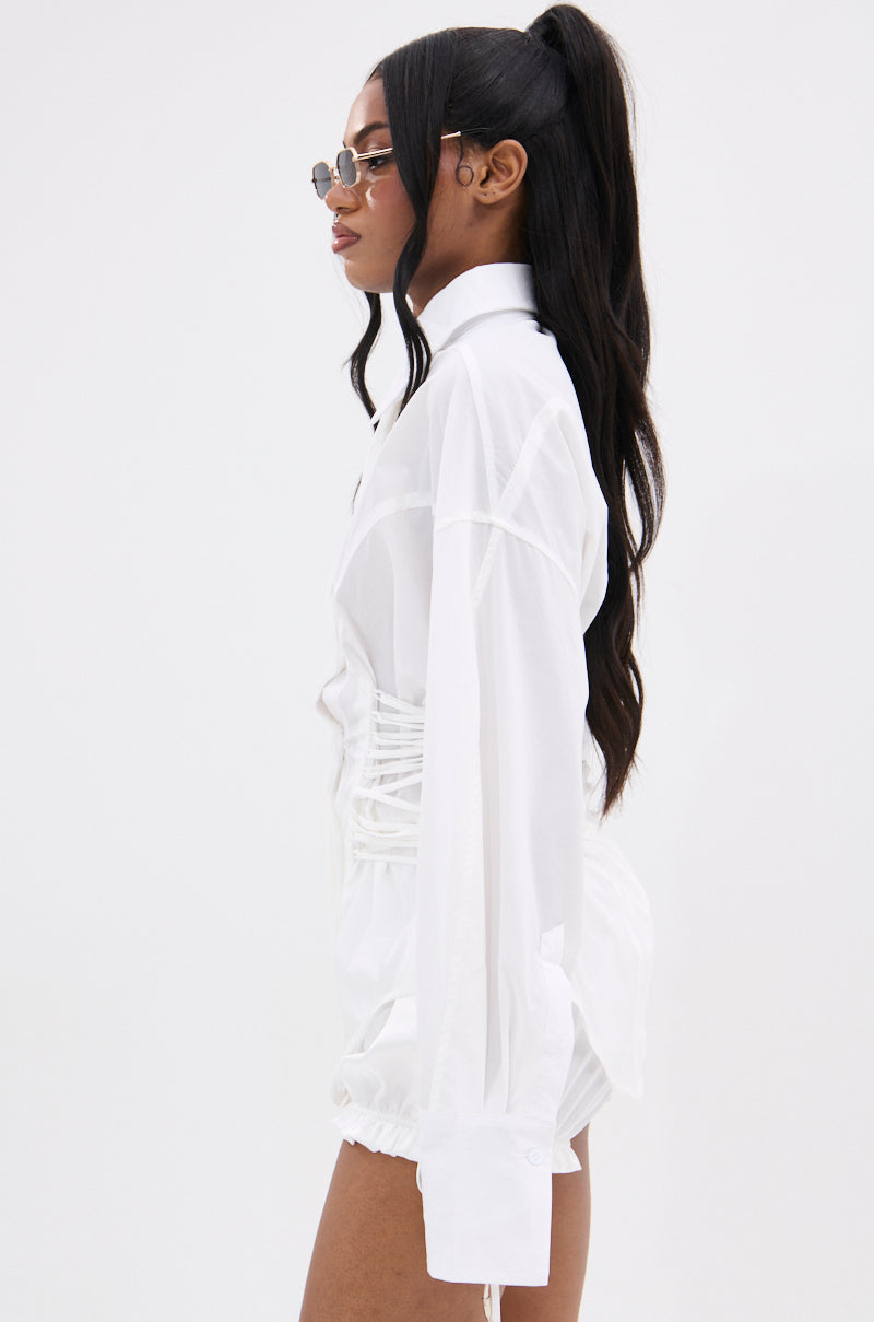 Woman in profile wears AKIRA Label sunglasses and the SHAR BUTTON DOWN WITH SIDE TIES, a long-sleeved white shirt with ruched sides and corset-inspired shape, paired with white shorts. Her long dark hair is styled in a high ponytail against a white background.