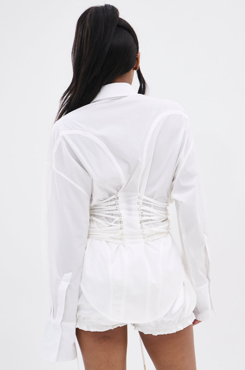 A person with long dark hair wears the SHAR BUTTON DOWN WITH SIDE TIES, a white shirt with a corset-inspired silhouette and long sleeves, paired with white shorts against a plain white background.
