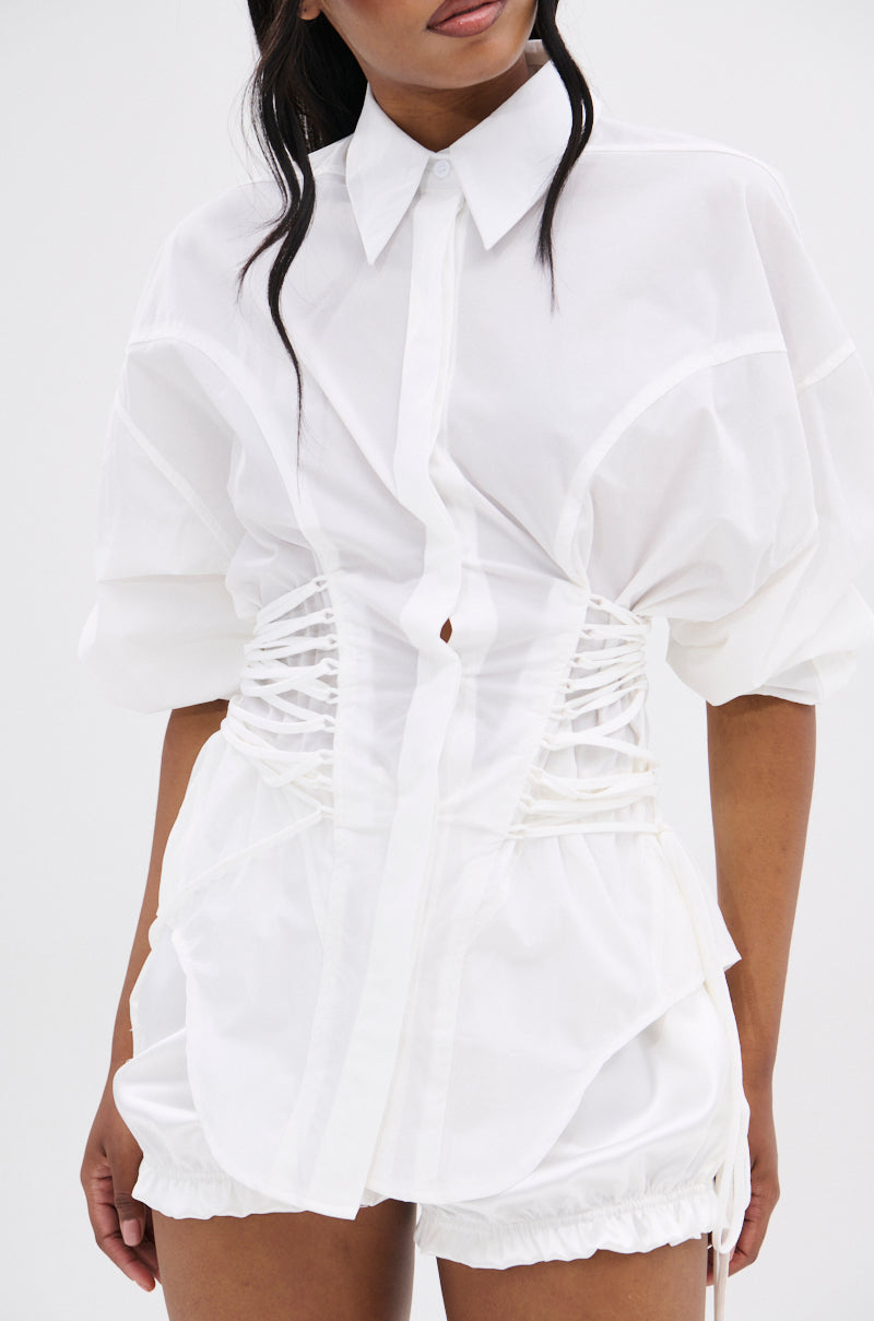She wears the SHAR BUTTON DOWN WITH SIDE TIES—a white blouse with a corset-inspired silhouette and side waist ties—paired with white shorts. Her hair falls in loose waves as she turns her head slightly.